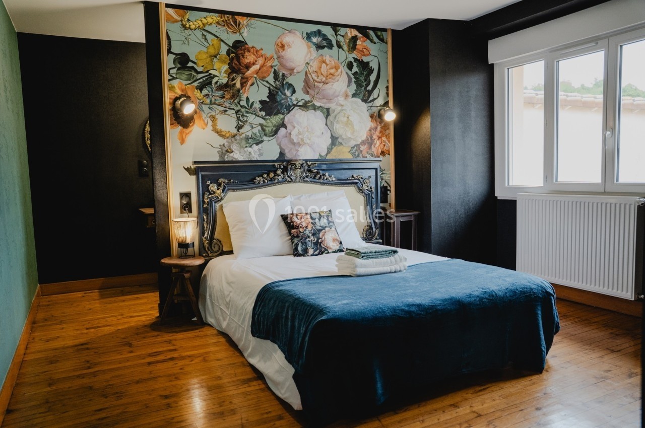 Chambre avec lit double, tête de lit ornée de fleurs, parquet en bois et grande fenêtre laissant entrer la lumière.