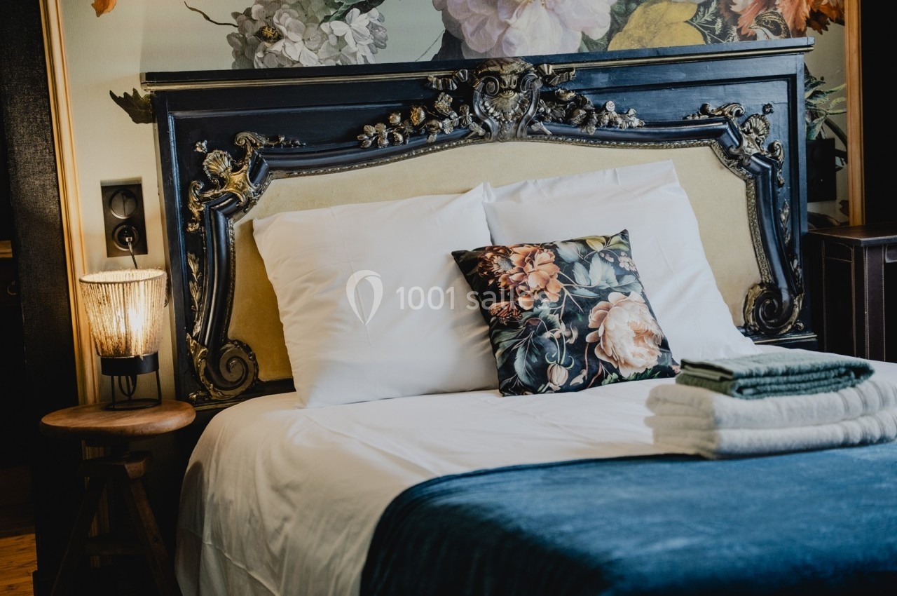 Lit avec tête de lit ornée, coussin à motif floral, lampe allumée sur une table en bois et serviettes pliées.
