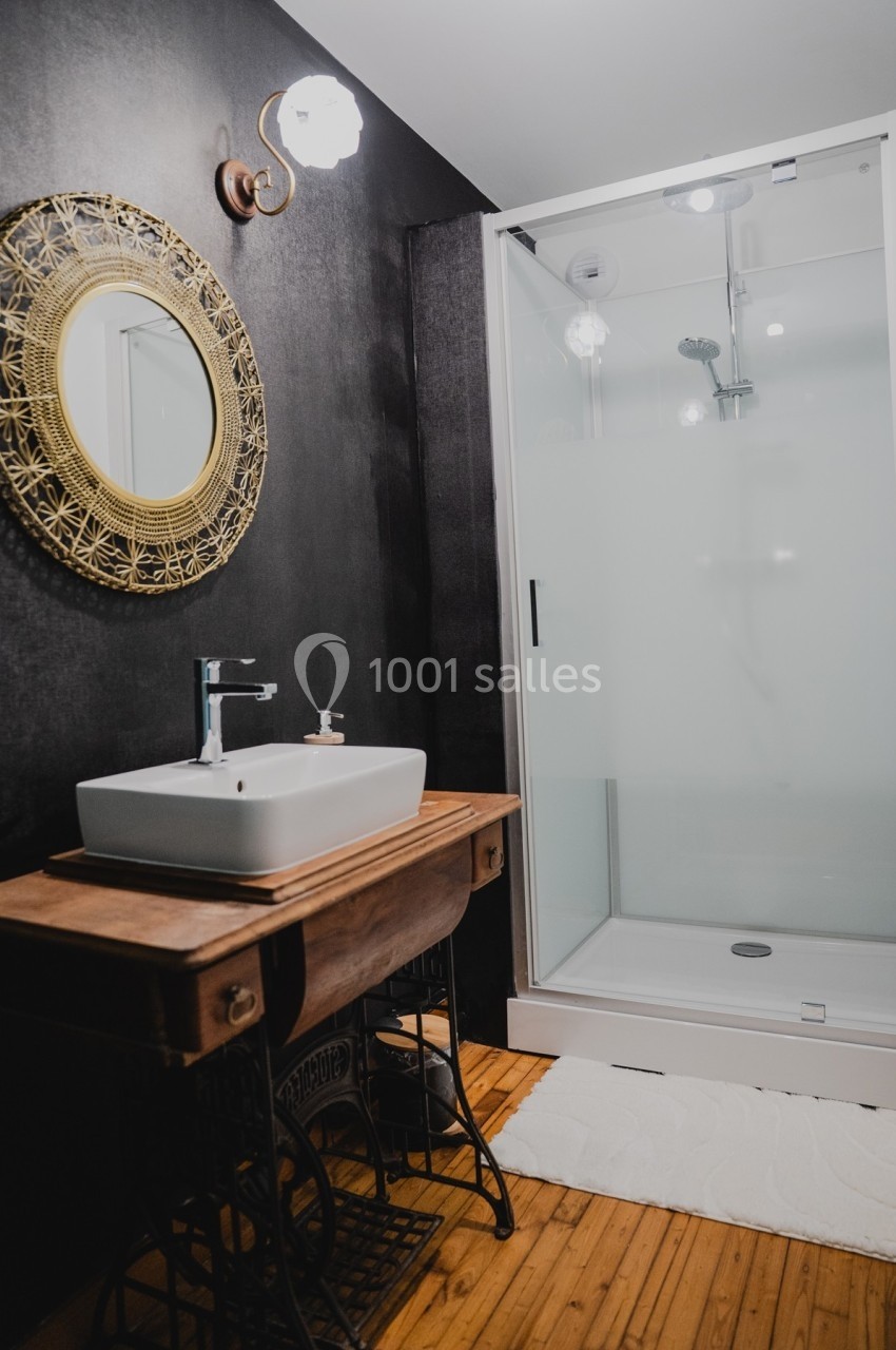 Salle de bain avec lavabo sur meuble en bois, miroir rond encadré et cabine de douche vitrée sur sol en bois.