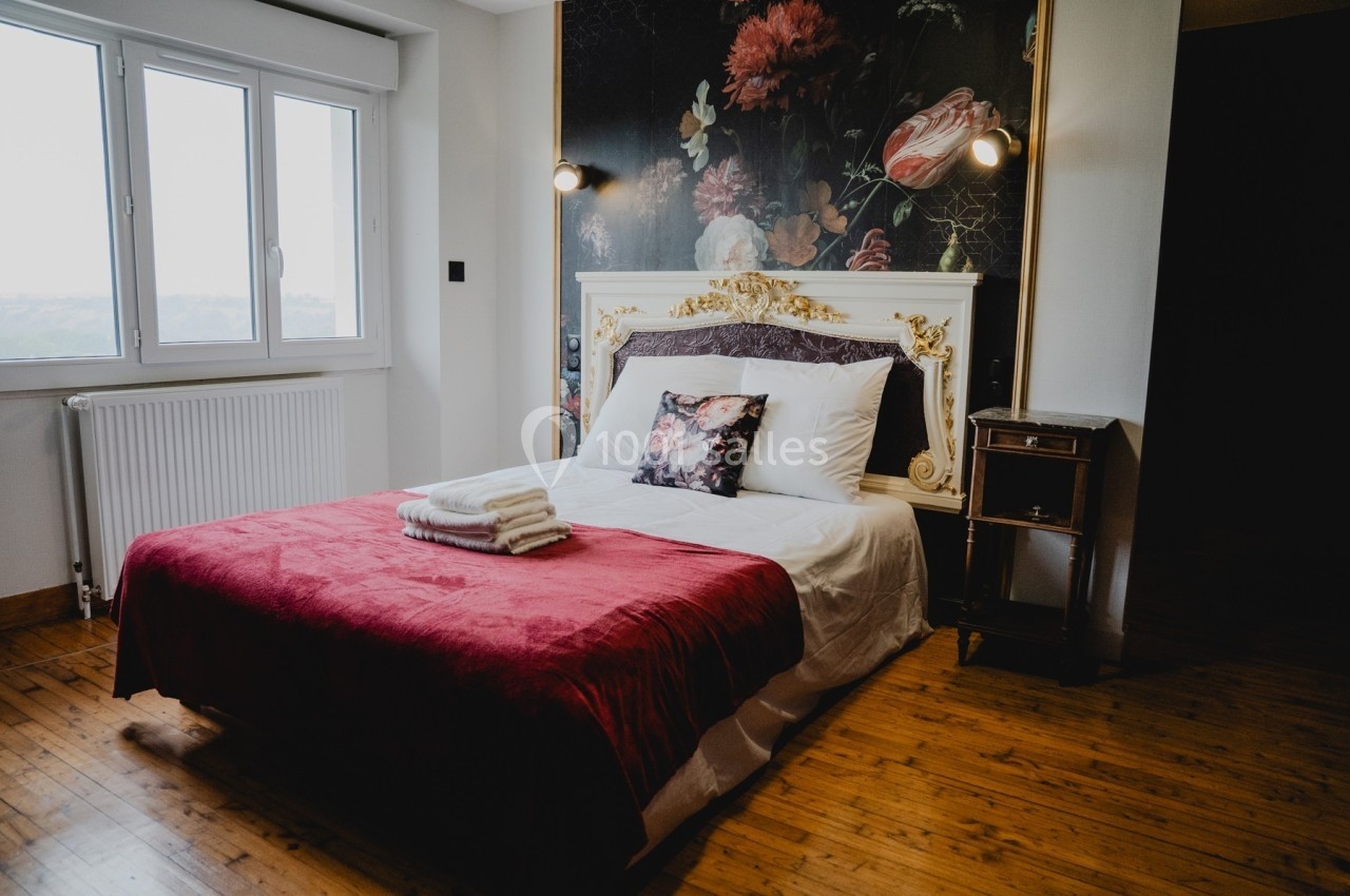 Lit double avec couverture rouge, oreillers décoratifs et serviettes pliées, dans une chambre lumineuse au parquet en bois.