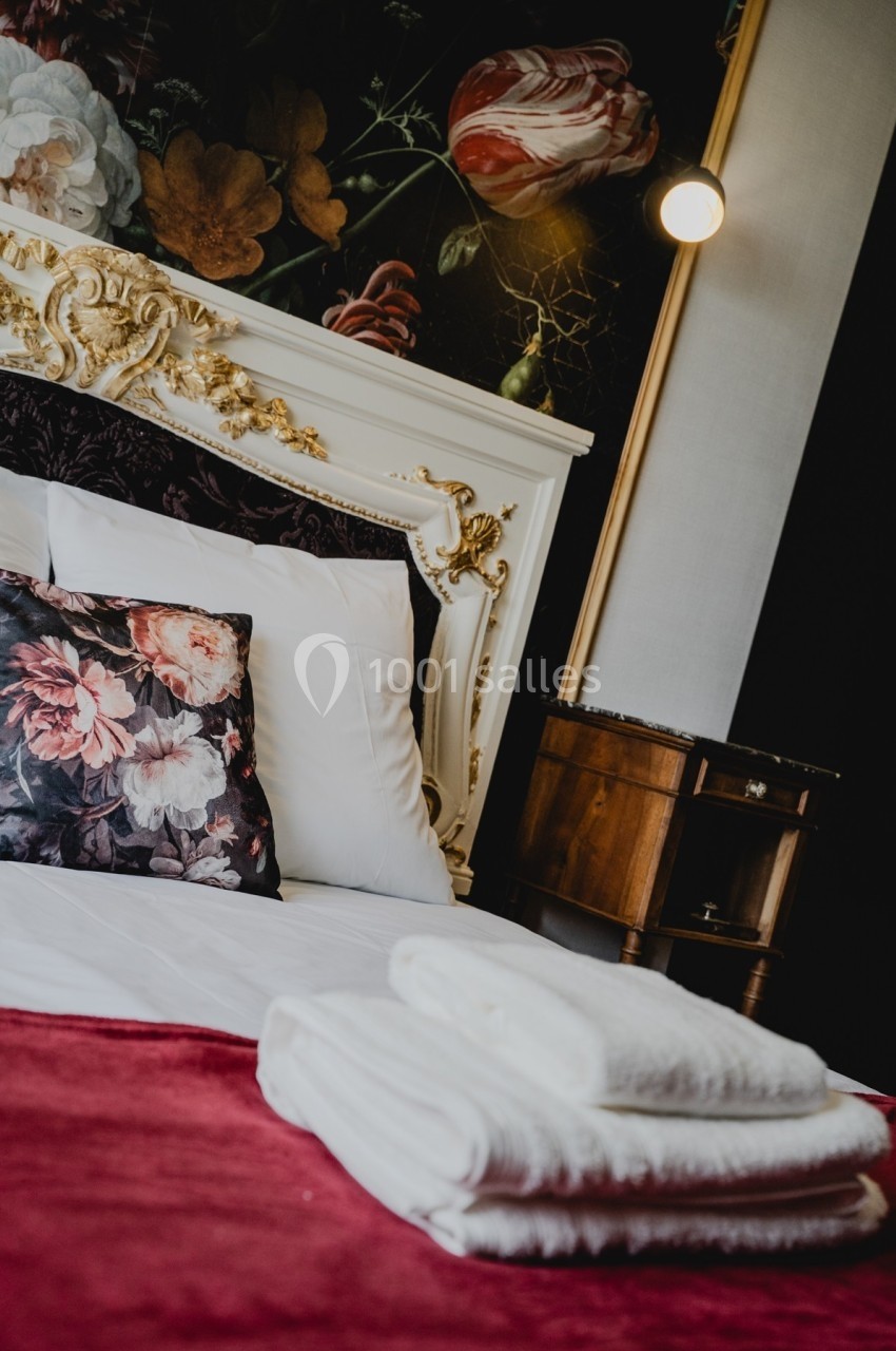 Lit avec tête de lit ornée de dorures, coussins fleuris, draps blancs et serviettes pliées sur une couverture rouge.