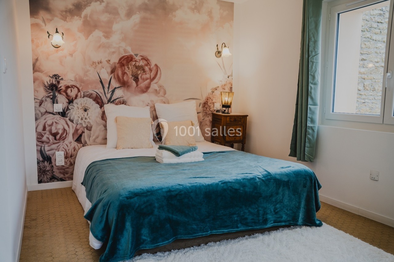Chambre avec un lit double, couvre-lit bleu, mur fleuri en arrière-plan, lampe de chevet et fenêtre à droite.