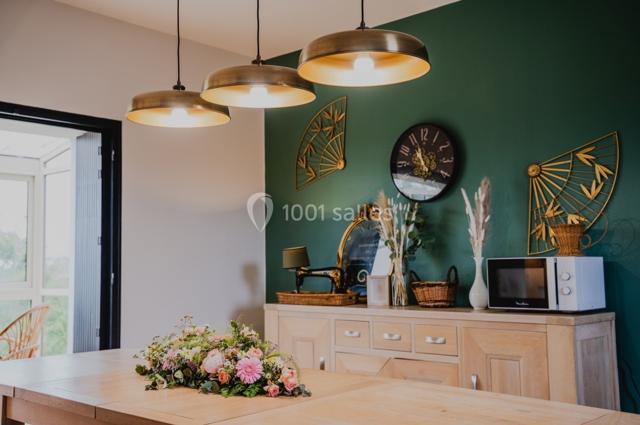Suspension lumineuse au-dessus d'un îlot central avec bouquet floral, mur vert décoré et mobilier en bois clair.