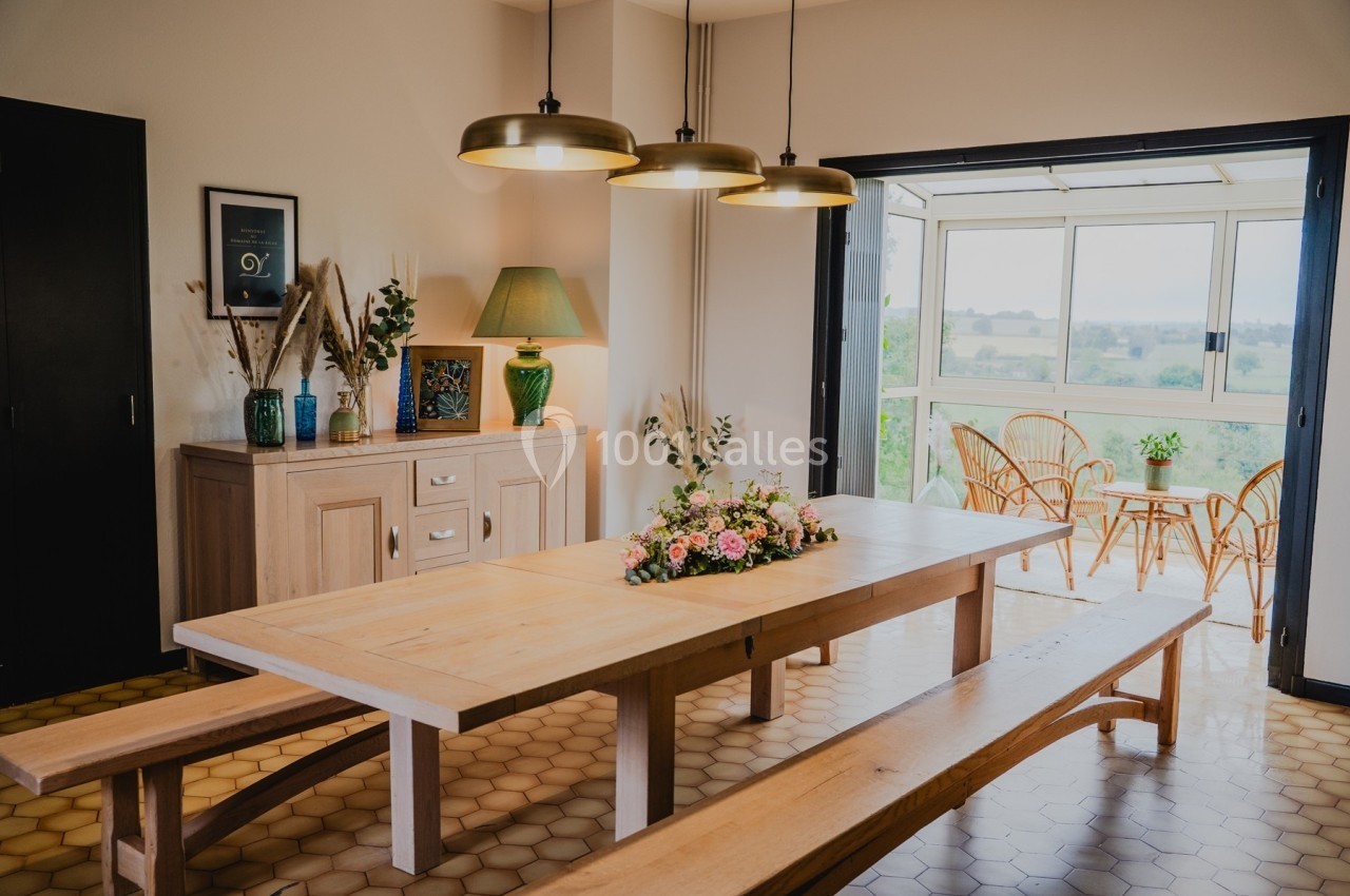 Salle à manger lumineuse avec une grande table en bois, des bancs, des luminaires suspendus et vue sur un balcon aménagé.