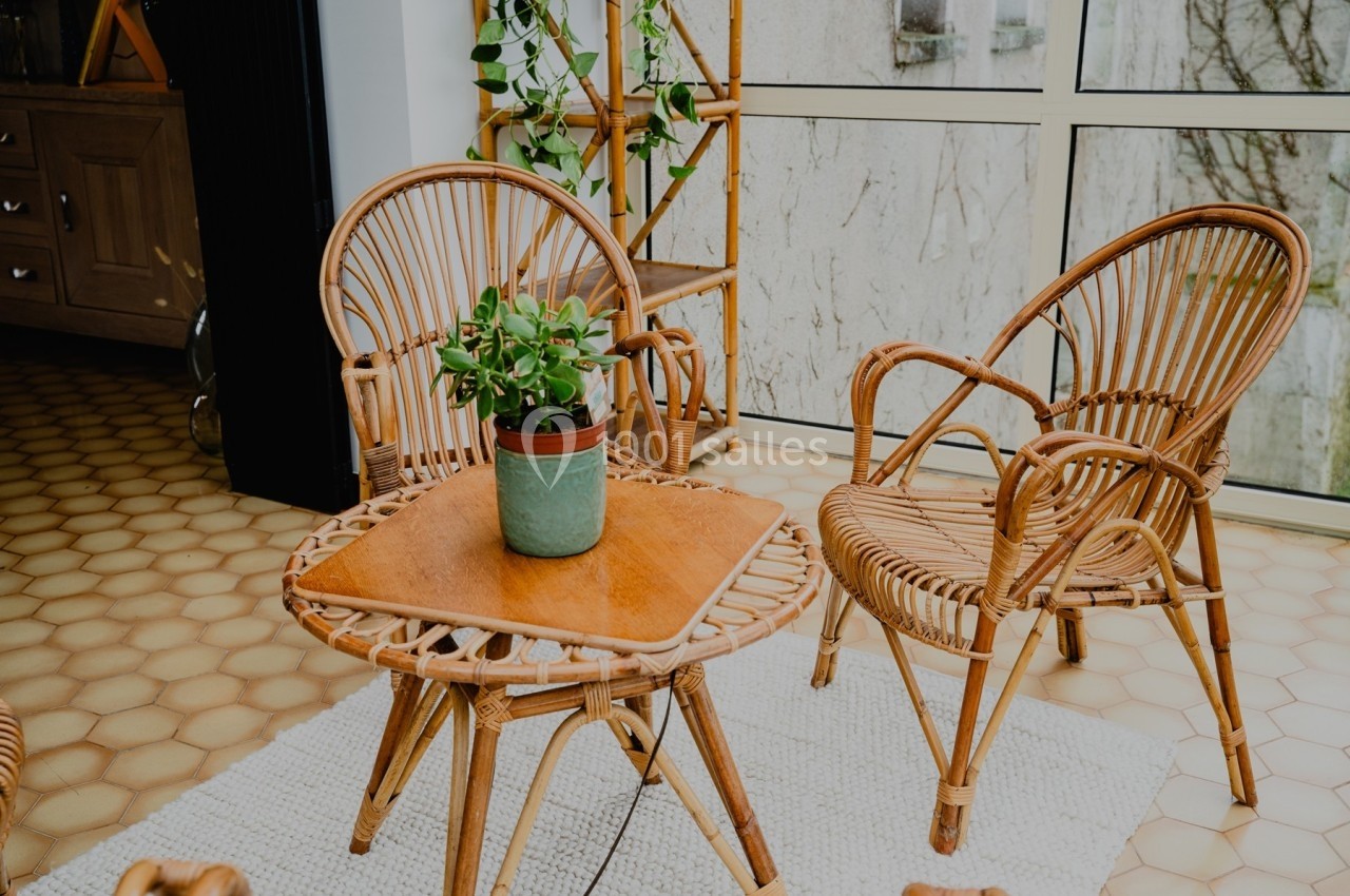 Deux fauteuils en rotin et une table assortie avec une plante en pot, dans une pièce lumineuse avec sol en carreaux.
