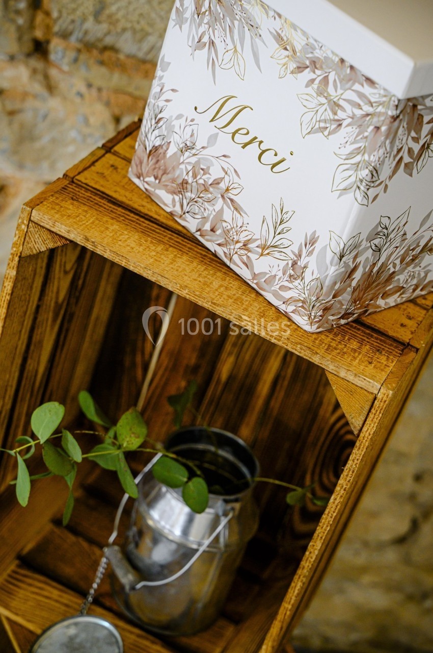 Boîte décorative avec inscription ’Merci’ posée sur une caisse en bois, à côté d'un arrosoir contenant des branches.