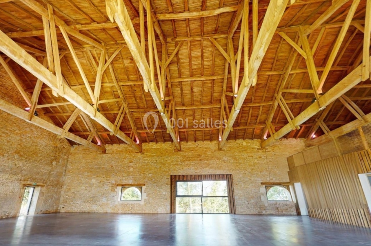 Charpente en bois apparente dans une grande salle aux murs en pierre, avec des fenêtres laissant entrer la lumière naturelle.