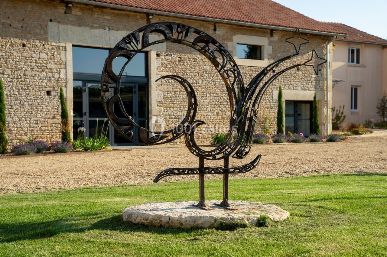 Sculpture métallique en forme de coq stylisé sur une pelouse devant un bâtiment en pierre avec des fenêtres modernes.