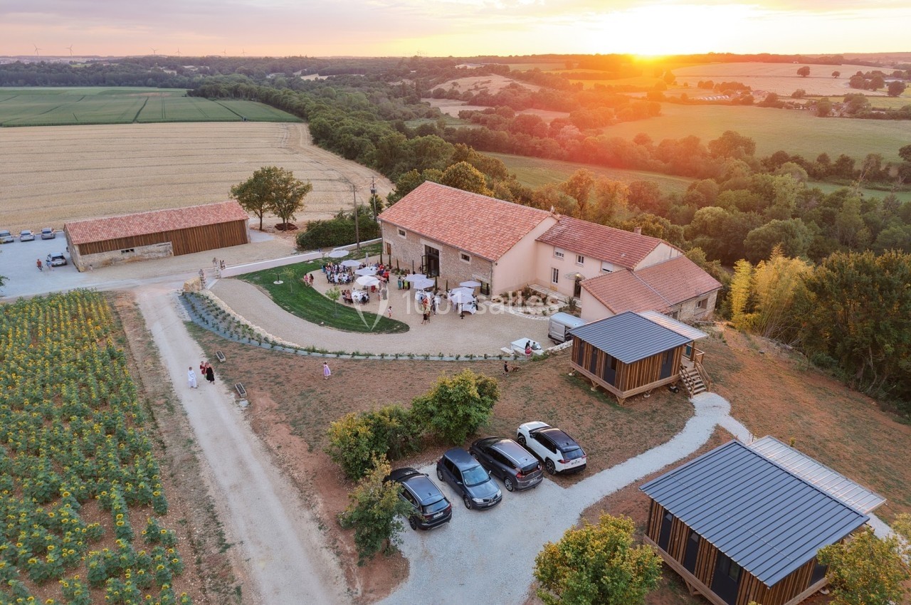 Vue aérienne d'une ferme rénovée avec des bâtiments en pierre, entourée de champs et d'un coucher de soleil.