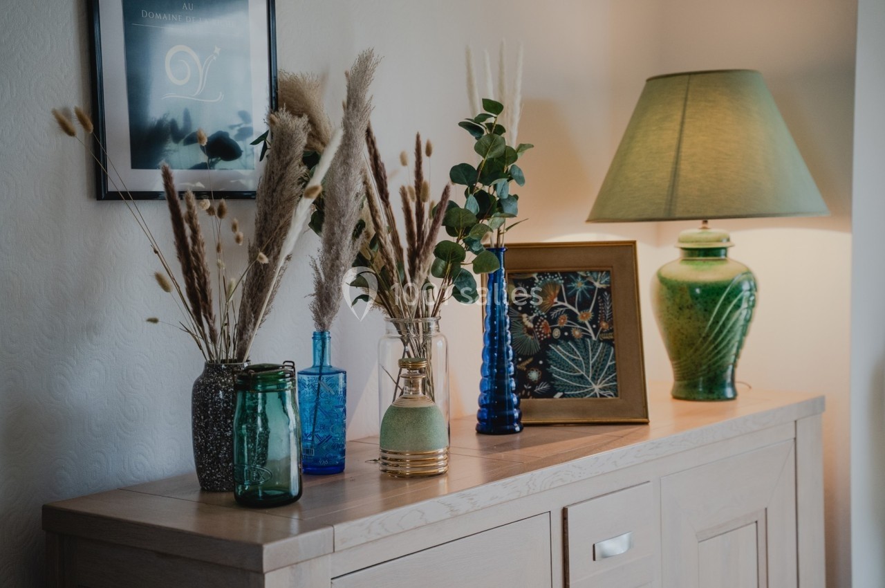 Composition de vases en verre coloré avec des fleurs séchées sur un buffet en bois, lampe verte et cadres en arrière-plan.