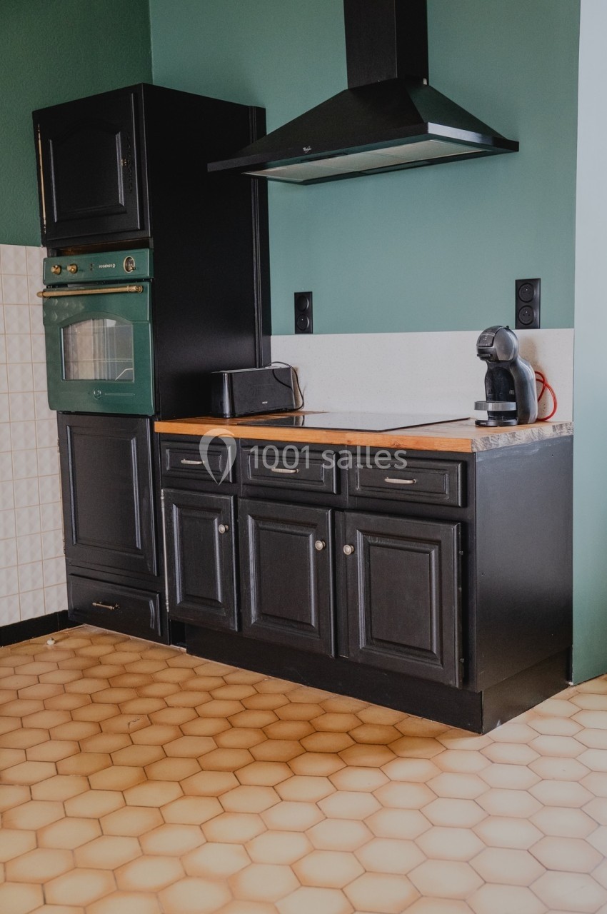 Cuisine avec meubles noirs, plan de travail en bois, hotte noire, four encastré et carrelage hexagonal beige au sol.