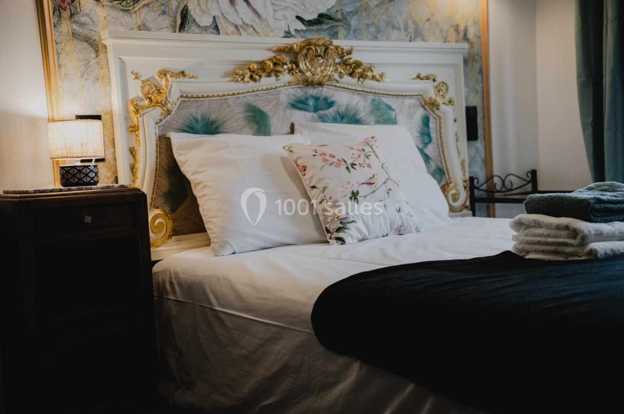 Lit avec tête de lit ornée de dorures, draps blancs, coussins décoratifs et lampe allumée sur une table de chevet.