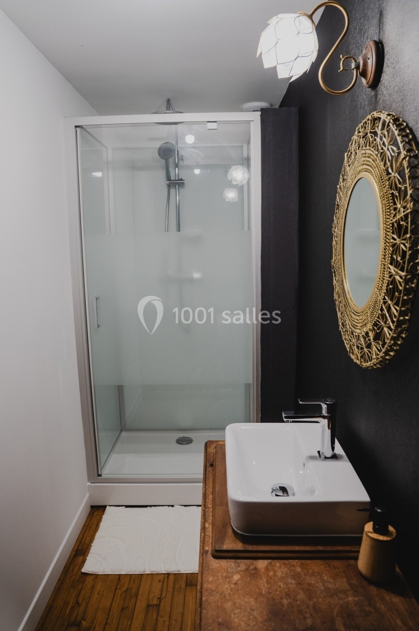 Salle de bain moderne avec douche vitrée, lavabo sur meuble en bois et miroir rond encadré doré.