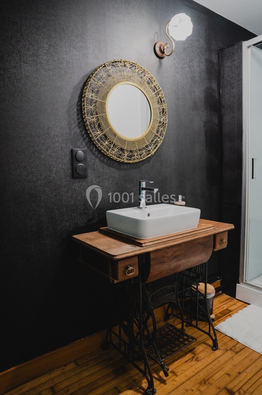 Lavabo moderne sur un meuble en bois avec miroir rond doré, mur noir texturé et sol en bois.