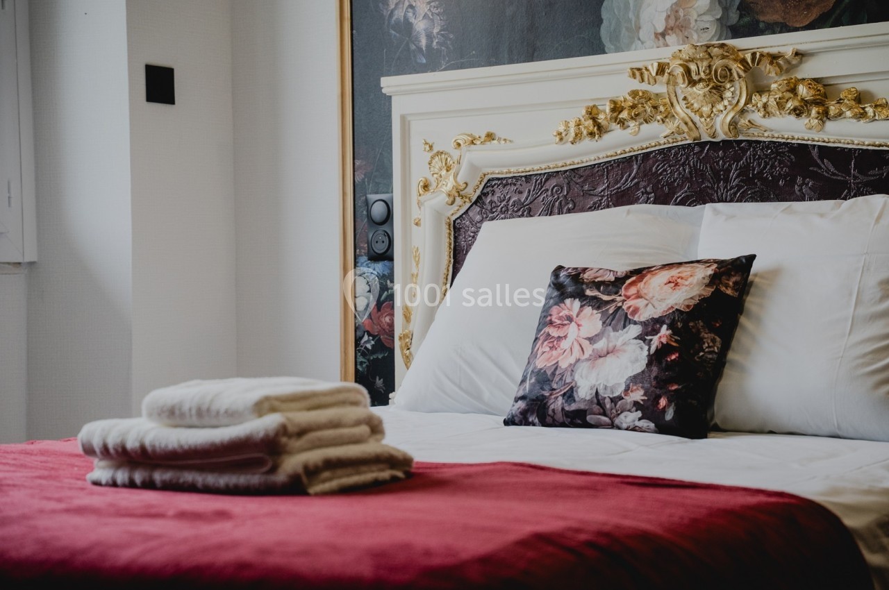 Lit avec tête de lit ornée, coussin à motifs floraux, couverture rouge et serviettes pliées posées dessus.