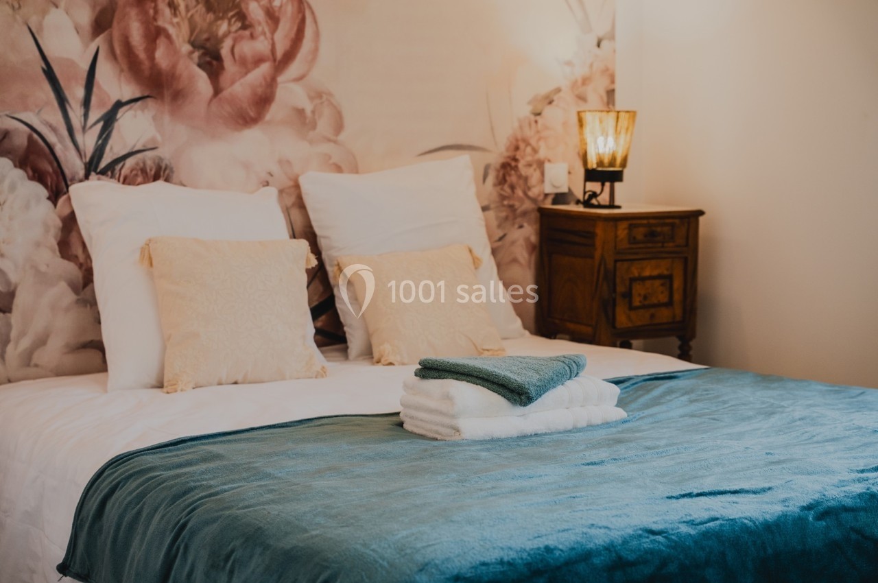 Lit double avec draps blancs, plaid bleu, coussins décoratifs et table de chevet en bois avec lampe allumée.