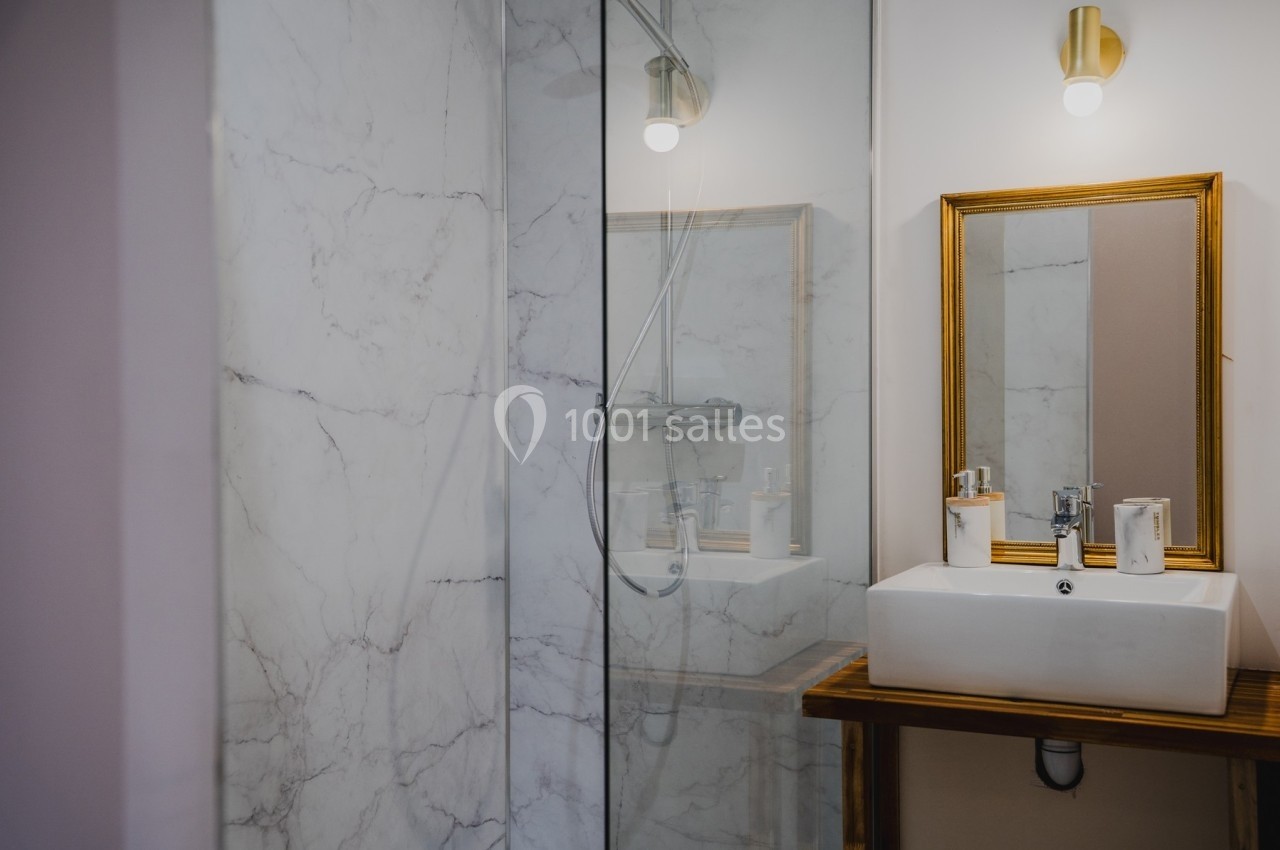 Salle de bain avec douche en verre, lavabo blanc sur meuble en bois et miroir encadré doré.