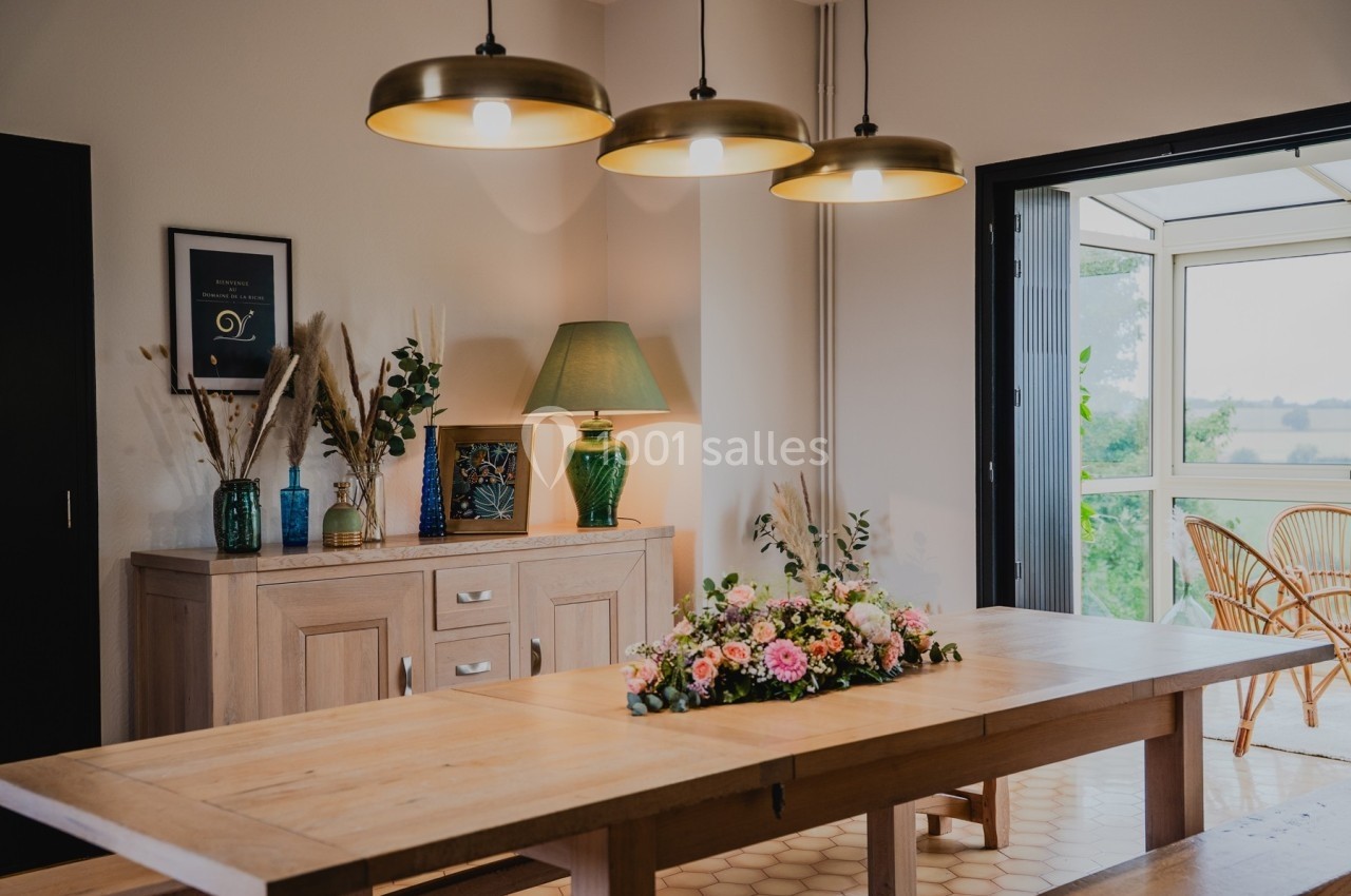 Salle à manger lumineuse avec une grande table en bois, décoration florale, buffet et suspensions dorées.