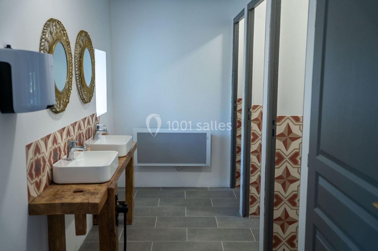Salle de bain avec deux lavabos sur un meuble en bois, miroirs ronds et cabines de toilettes au fond.