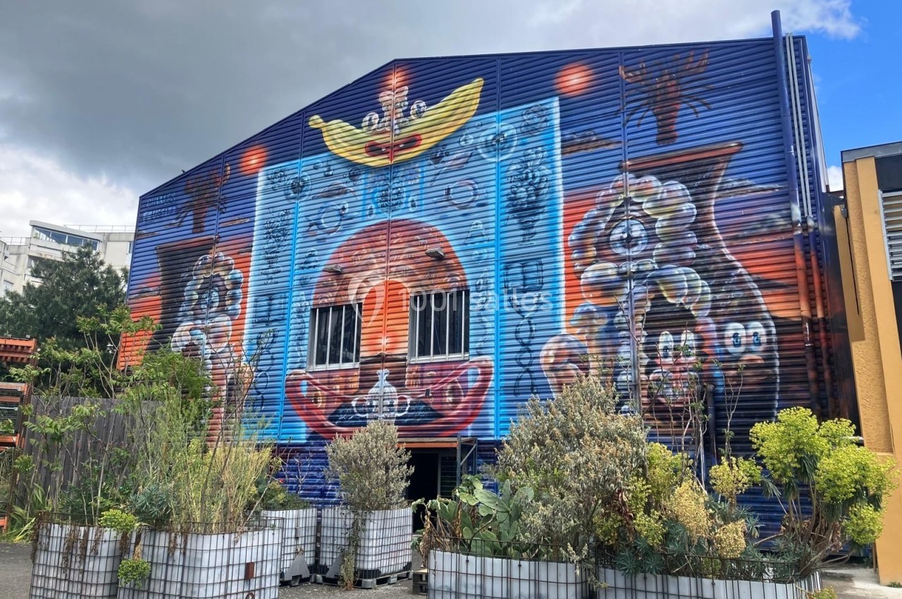 Façade d’un bâtiment industriel avec une fresque colorée et des plantes en bacs au premier plan.
