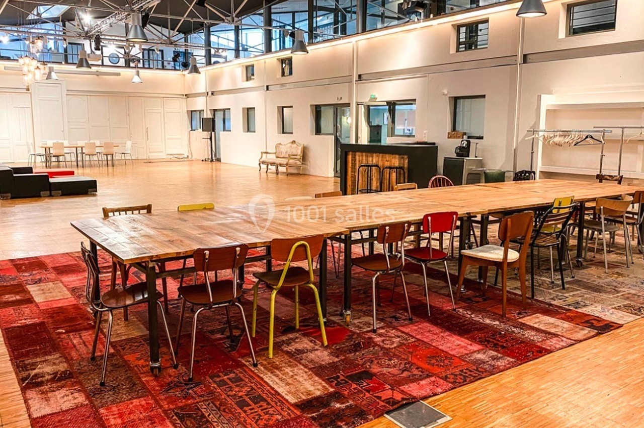 Grande salle lumineuse avec une longue table en bois entourée de chaises dépareillées, sur un tapis rouge à motifs.