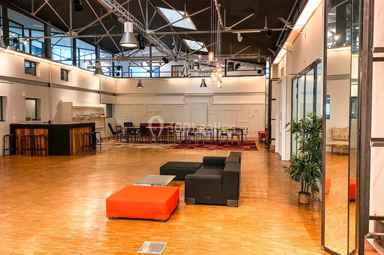 Salle spacieuse avec parquet, mobilier moderne, canapés, tables et éclairage industriel sous une verrière.