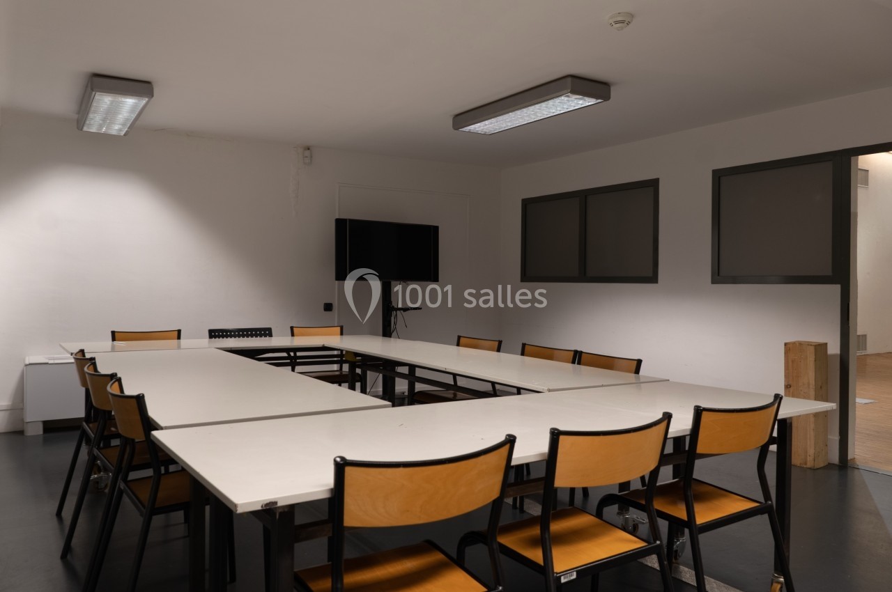 Salle de réunion vide avec une grande table en U, des chaises en bois et un écran noir fixé au mur.