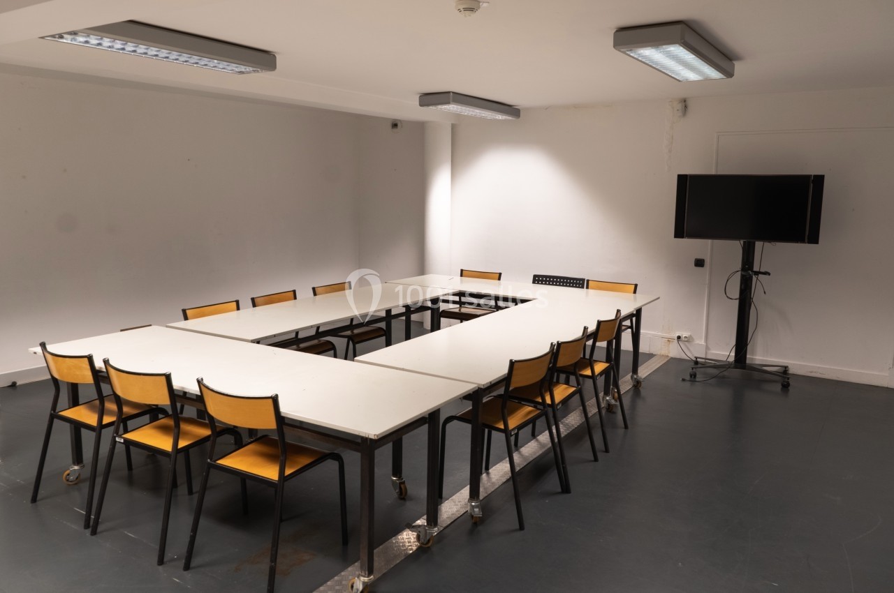 Salle de réunion vide avec des tables disposées en U, des chaises en bois et un écran noir sur pied.
