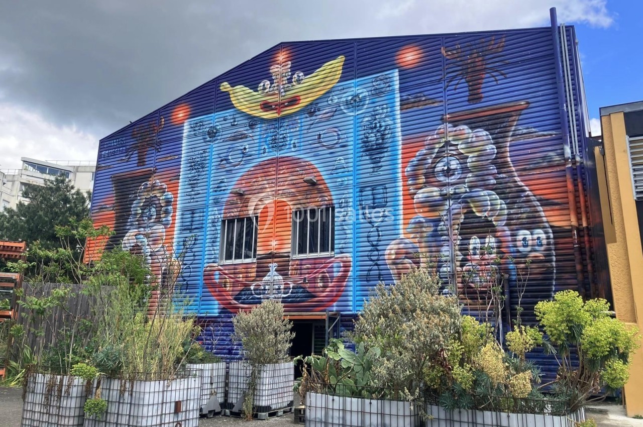 Fresque colorée sur un bâtiment industriel, représentant des motifs abstraits et une barque, entourée de végétation.
