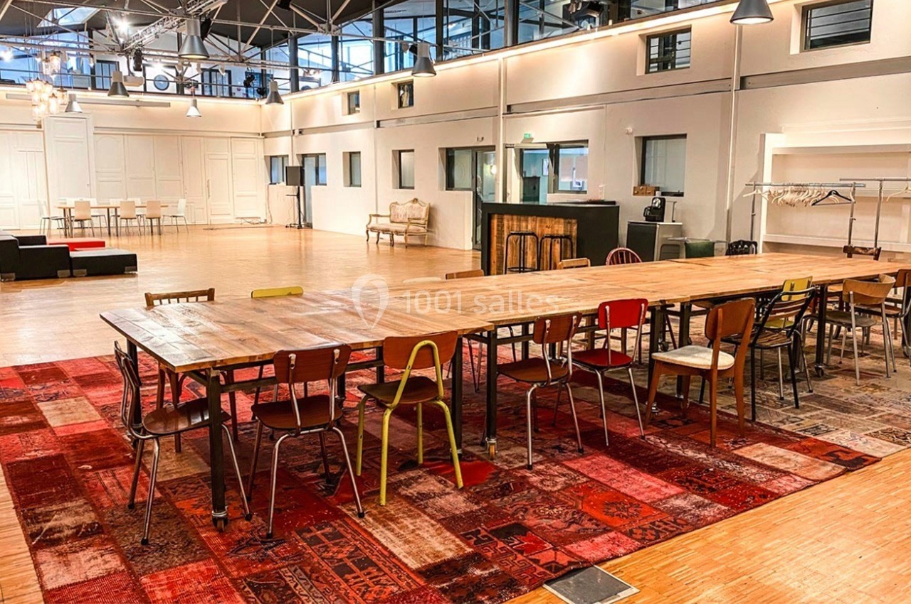 Grande salle lumineuse avec une longue table en bois entourée de chaises dépareillées, sur un tapis rouge à motifs.