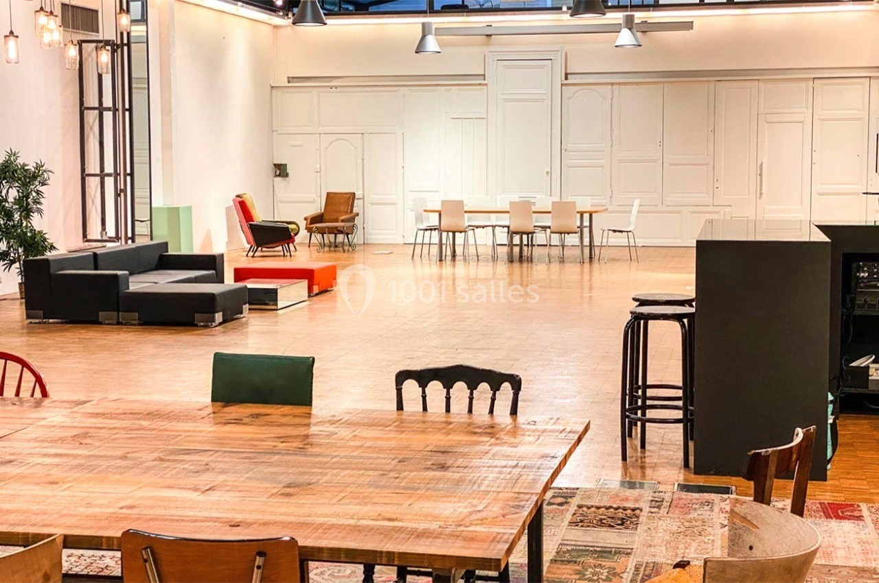 Salle spacieuse avec parquet, mobilier varié, tables en bois et chaises, éclairée par des lampes suspendues.