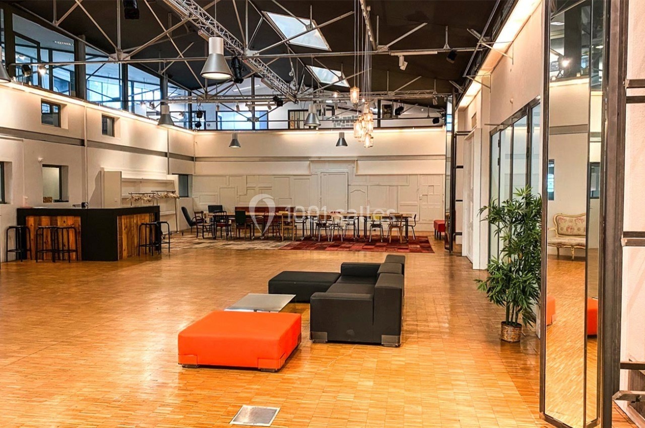 Salle spacieuse avec parquet, mobilier moderne, canapés colorés et éclairage industriel sous une structure métallique.