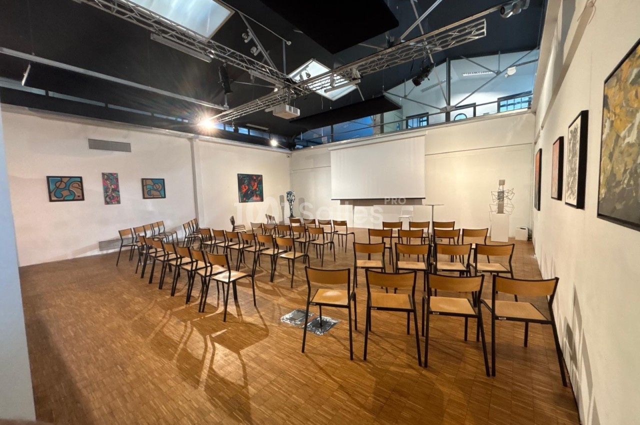 Salle lumineuse avec des chaises disposées en rangées face à un écran de projection, murs ornés de peintures.