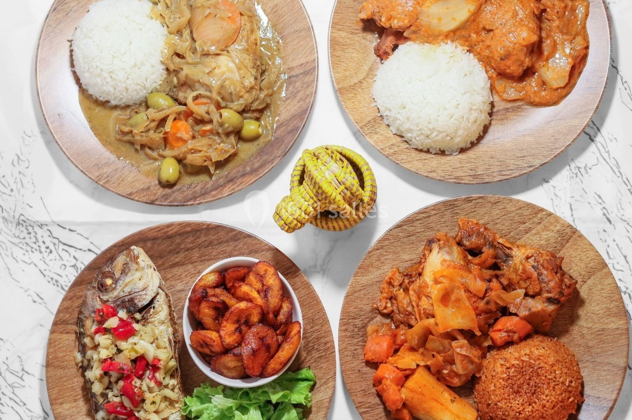 Quatre assiettes de plats africains variés avec riz, poisson, bananes plantains, légumes et sauce, disposées sur une table.