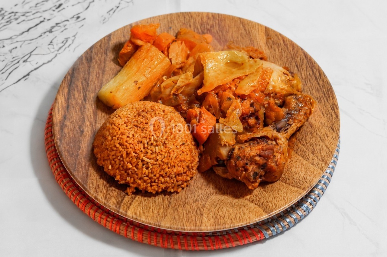 Assiette en bois contenant du poulet rôti, légumes mijotés et boule de couscous rouge, posée sur une nappe blanche.