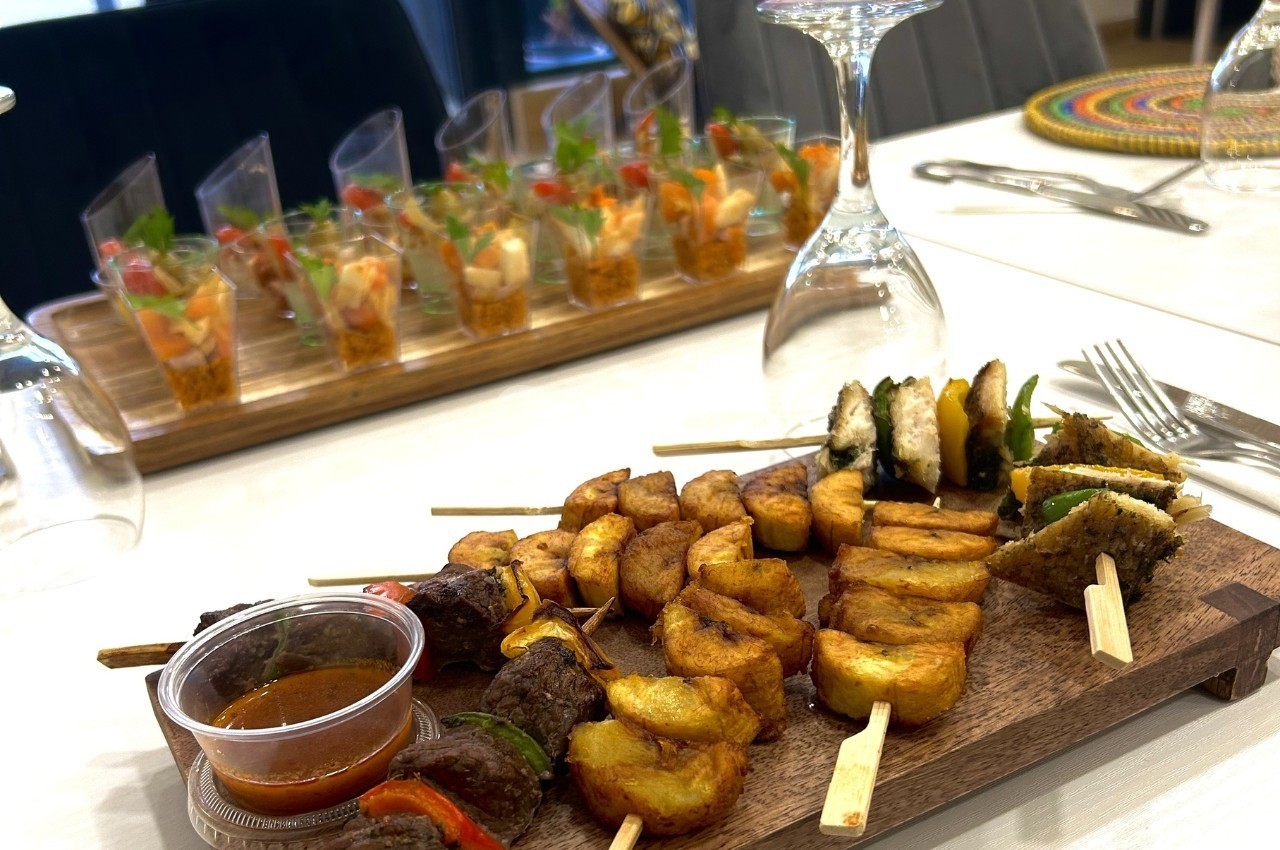 Plateau de brochettes de viande, bananes plantains et sauce, avec verrines apéritives en arrière-plan.