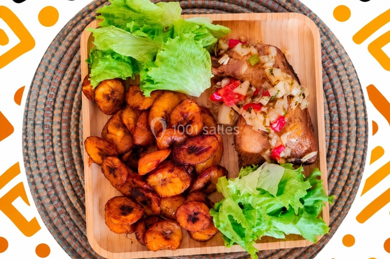 Assiette en bois contenant des rondelles de bananes plantains frites, du poulet garni de légumes et des feuilles de salade.