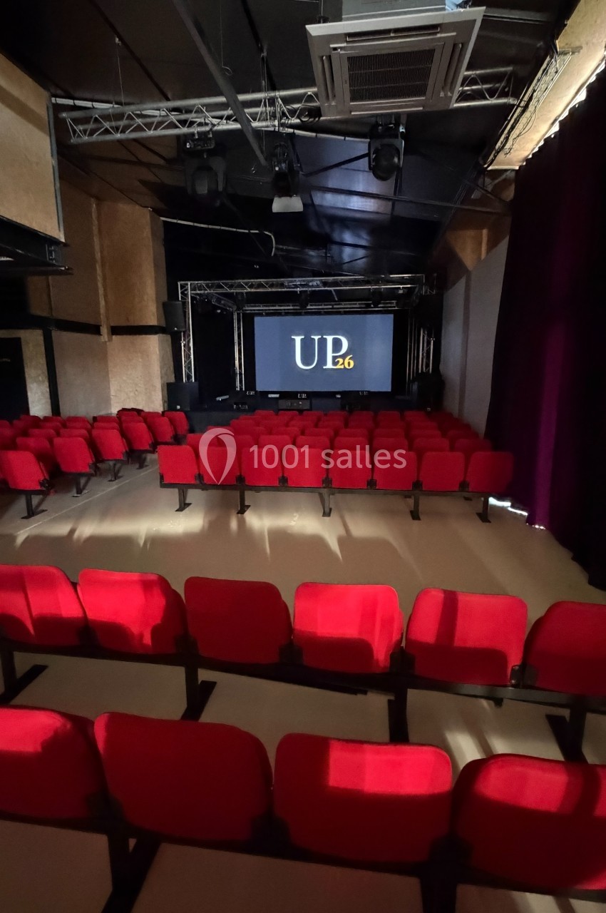 Salle de projection avec rangées de sièges rouges et un écran affichant ’UP26’ dans un espace sombre.