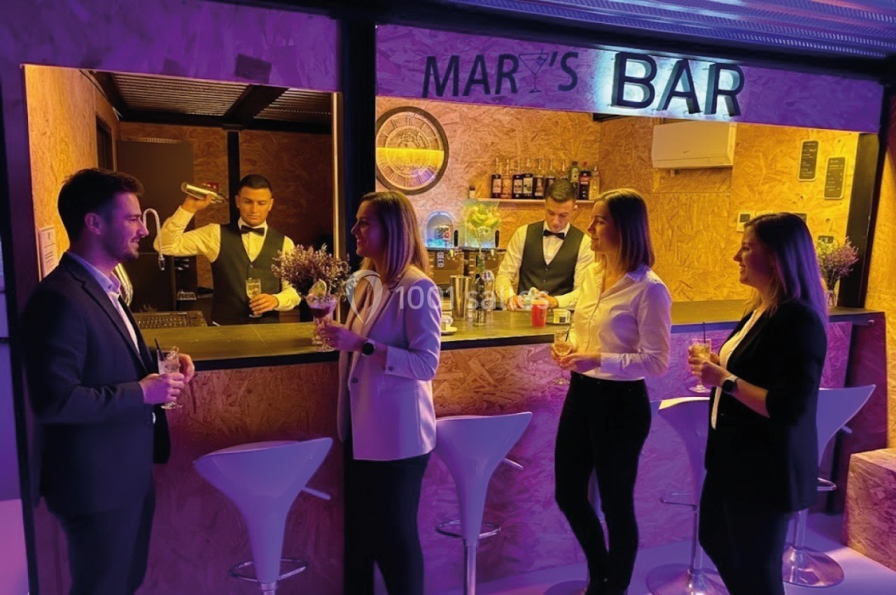 Groupe de personnes discutant et tenant des verres devant un bar éclairé, avec un barman en arrière-plan.