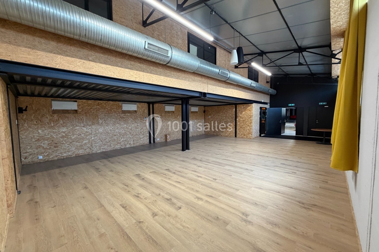 Salle spacieuse avec parquet clair, murs en bois, plafond industriel avec conduits apparents et mezzanine métallique.