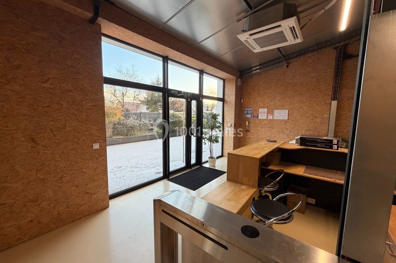Entrée d'un bureau avec comptoir en bois, chaise, baie vitrée donnant sur un extérieur arboré et sol clair.