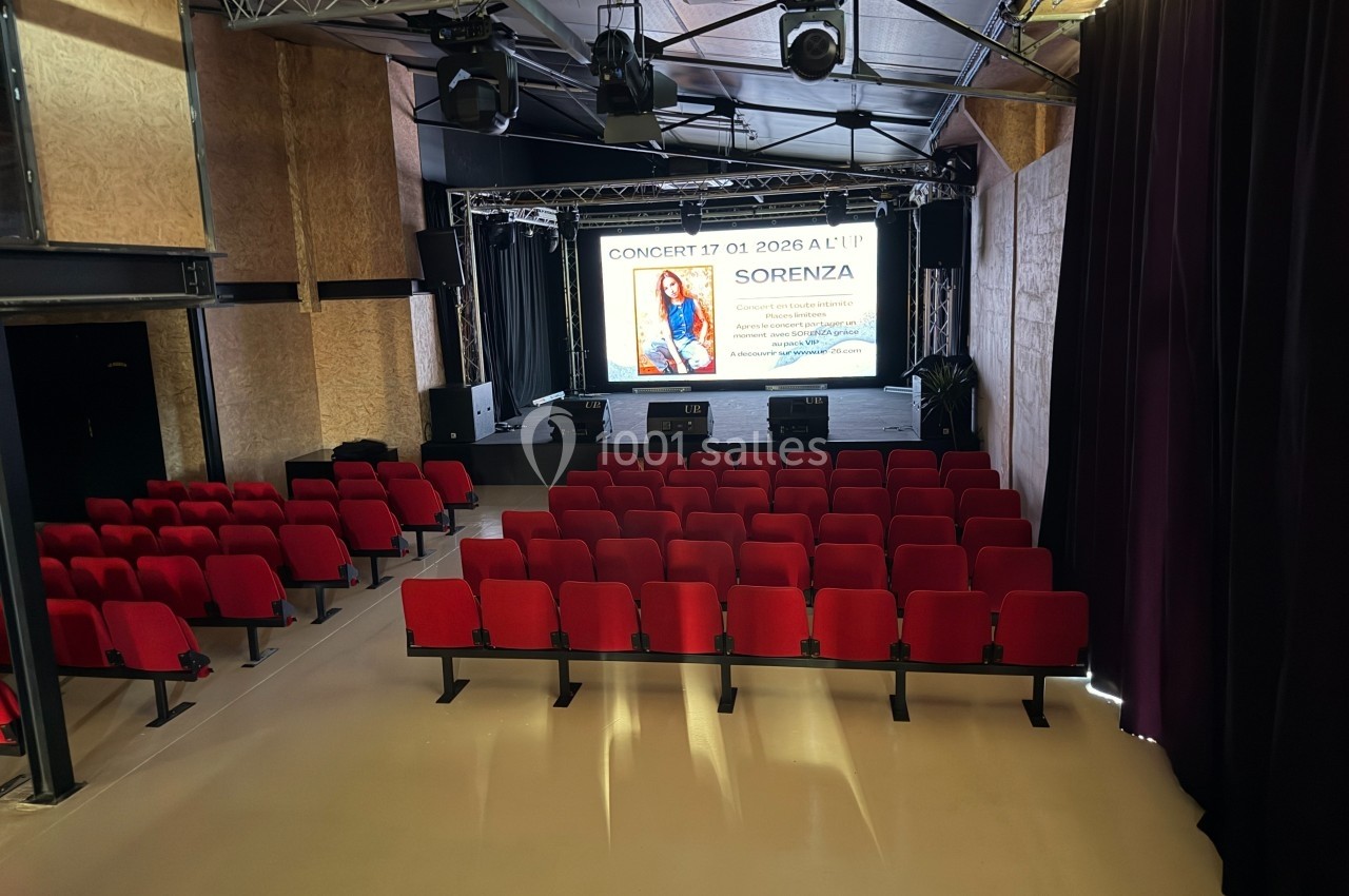 Salle de spectacle avec des rangées de sièges rouges face à une scène équipée d'un écran affichant une affiche de concert.
