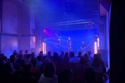 Salle de spectacle avec scène éclairée, artistes en représentation et public assis dans des fauteuils rouges.