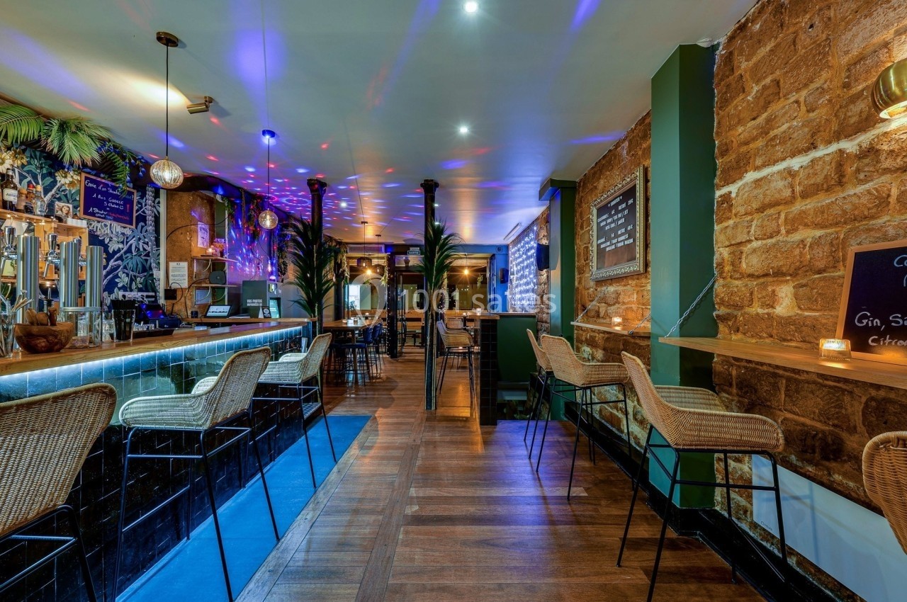 Intérieur d'un bar avec comptoir en bois, tabourets en rotin, éclairage tamisé et décorations végétales.