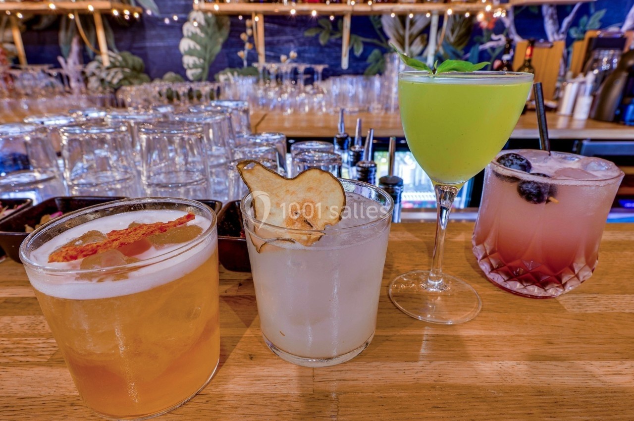 Quatre cocktails variés alignés sur un comptoir en bois, avec des garnitures décoratives et un bar en arrière-plan.