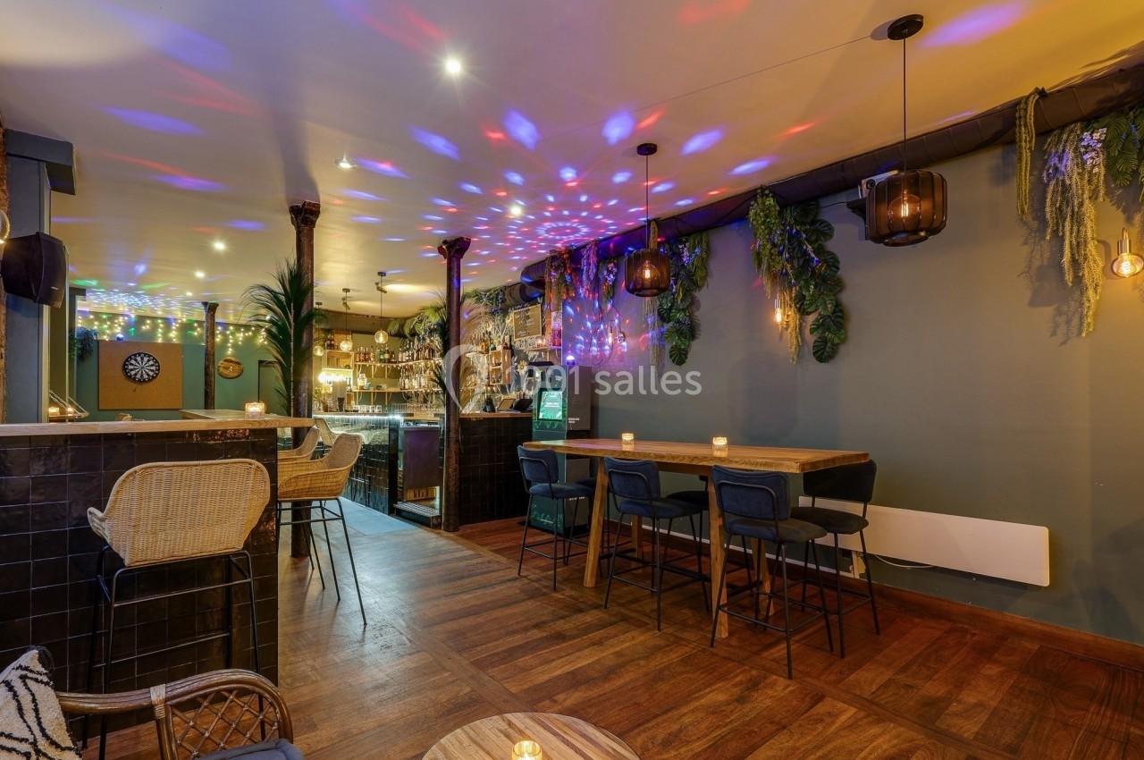 Intérieur d'un bar chaleureux avec éclairage tamisé, décor végétal, tables en bois et lumières colorées au plafond.