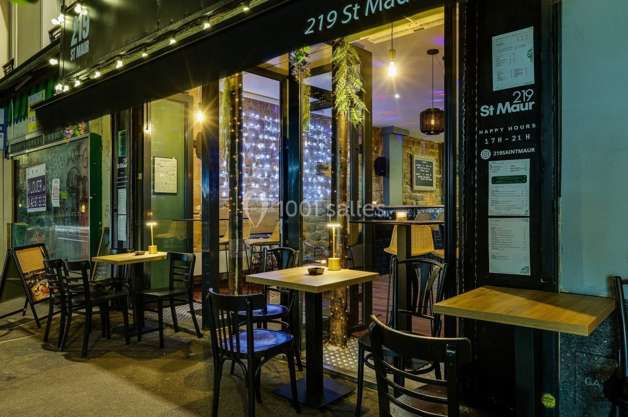 Façade d'un café-restaurant avec terrasse éclairée, tables et chaises en extérieur, ambiance chaleureuse en soirée.