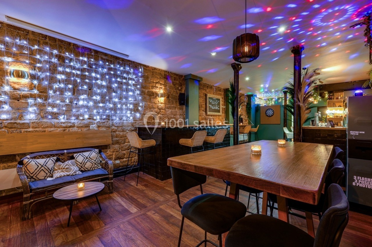 Salle de bar chaleureuse avec murs en pierre, guirlandes lumineuses, table en bois, tabourets et espace détente.