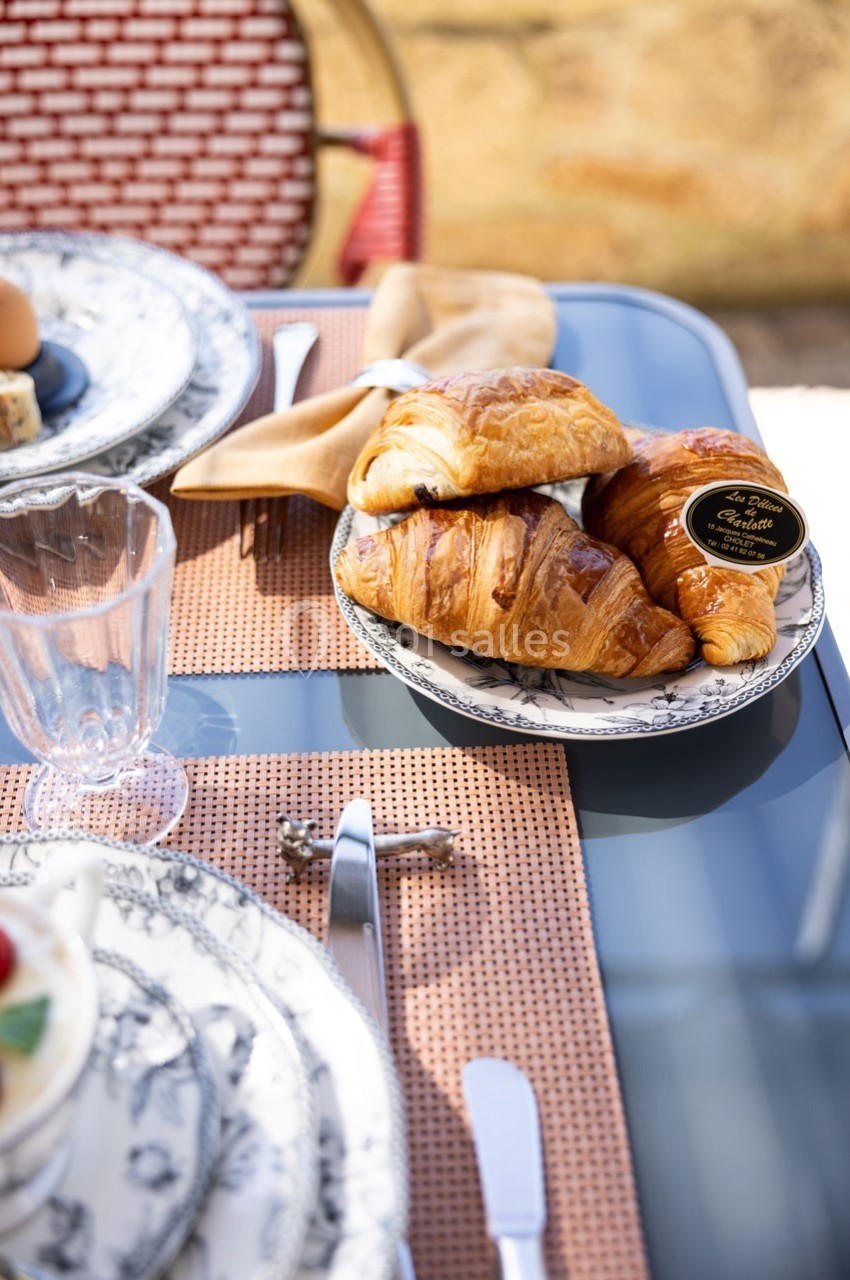 Assiette de croissants sur une table dressée en extérieur avec des couverts, un verre et une nappe tressée.