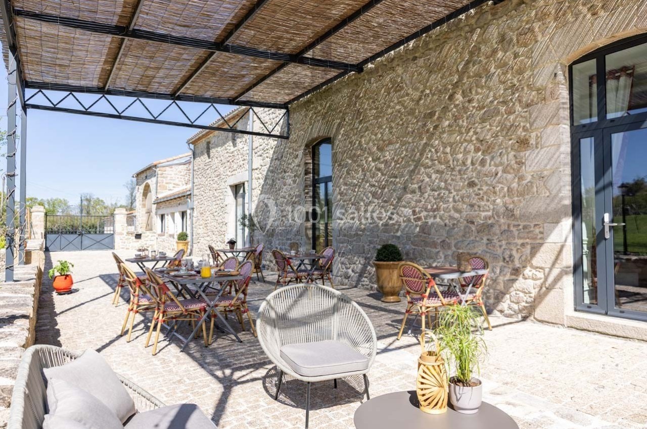 Terrasse ensoleillée avec mobilier de jardin, tables dressées, et façade en pierre sous une pergola en bois.